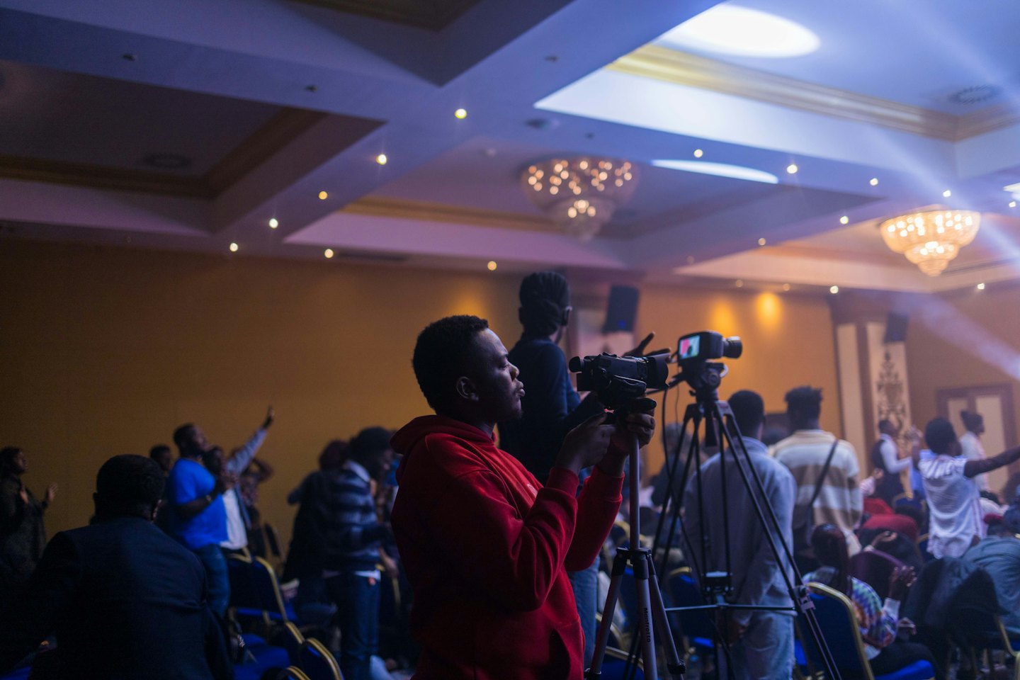 Videographer records event with diverse attendees in dimly lit venue.